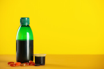 Bottle of syrup, measuring cup and cough drops on yellow background. Space for text