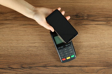 Woman with smartphone using modern payment terminal at wooden table, top view