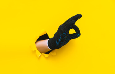 OK sign of two connected fingers in the form of a ring in black knitted work gloves on a yellow background with a torn paper hole and copy space. The sign is excellent.