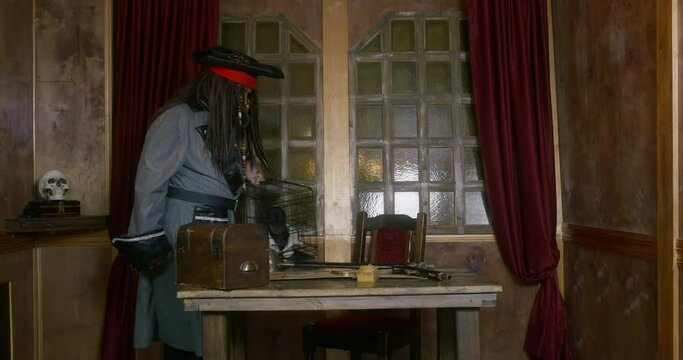 Pirate captain with long hair and coat puts iron cage with dachshund ON table near revolver against window in cabin of ship