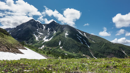 A picturesque mountain range against the blue sky. Patches of snow are visible on the slopes. Lush green grass and wildflowers grow in the valley, in the meadow. Kamchatka. Vachkazhets 