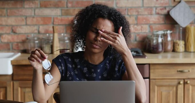 Tired young Black female exhausted with online work on laptop suffer of eye strain take glasses off rub nose bridge. Fatigue millennial woman remote employee feel pain in eyes overworked by computer