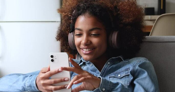 Cute curly haired African teenage girl wear wireless headphones wave hand greeting friend start videoconference use cellphone, close up. Young generation modern tech usage, video call event concept
