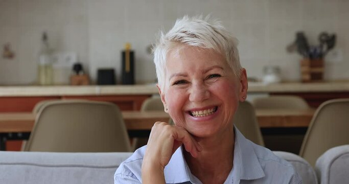 Head shot close up portrait happy 50s woman sit on sofa pose at camera, having wide toothy charming smile enjoy carefree retired life relax at home. Medical insurance cover for older citizen concept