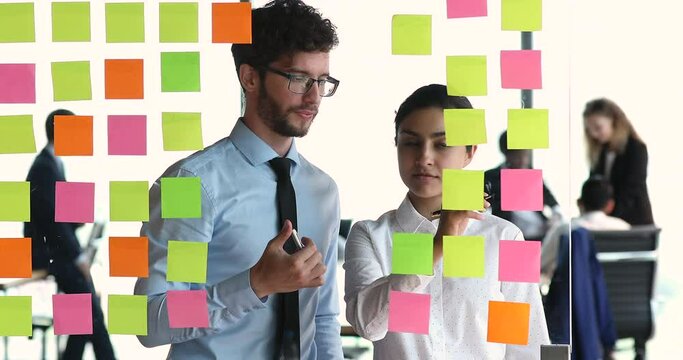Young coworkers program code writers create software interface structure place colored post it paper sheets on glass wall. Two mates managers Indian woman European man talk make marks on kanban cards