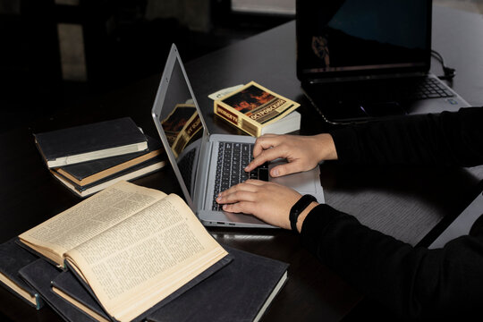 There are books and a computer on the table. Male or female hands work on a laptop. A writer or content creator writes text for a magazine, website, or advertisement.