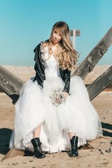 Blonde girl in a wedding dress and a leather jacket in a rock style on a wild sandy beach