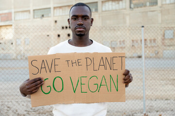 Young African American man looking at camera holding cardboard sign Save the planet, go vegan