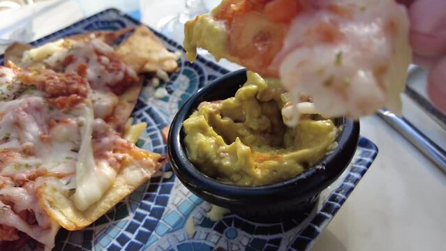 Dipping a nacho with cheese in a small bowl with guacamole. A beautiful blue plate with nachos and cheese on a table outdoors. Hand dipping a piece of a nacho chip in guacamole.