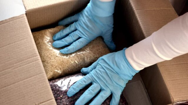 An online order packing operator in blue rubber gloves puts packages of different cereals into a box: oatmeal, beans, rice. The concept of food delivery or charity. View from above.