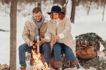Naklejka na ściany i meble A couple in love, a man and a woman in a winter forest around a campfire drink coffee and give marshmallows