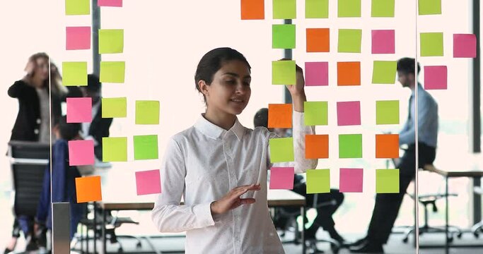 Smiling Indian businesswoman project manager write compare data on colored stickers based on glass board. Young lady software engineer work with information develop new task using kanban or scrum tool
