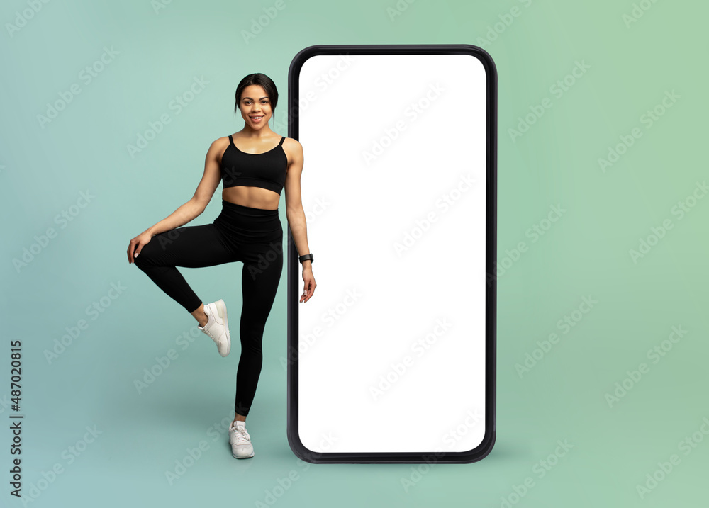 Wall mural mobile app for online workout. young fitness lady standing near huge smartphone with white blank scr