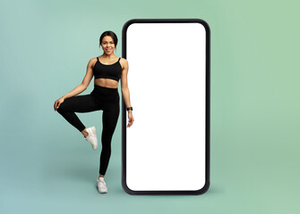 Mobile app for online workout. Young fitness lady standing near huge smartphone with white blank screen, mockup