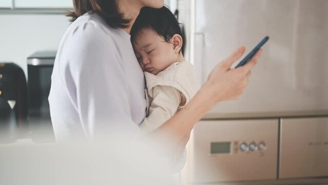 眠る赤ちゃんを抱っこしながらスマートフォン操作をするお母さん