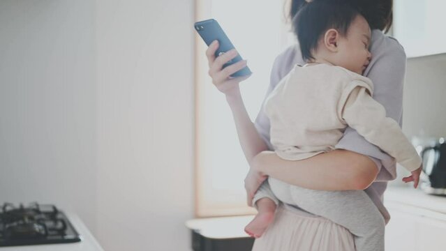 眠る赤ちゃんを抱っこしながらスマートフォン操作をするお母さん