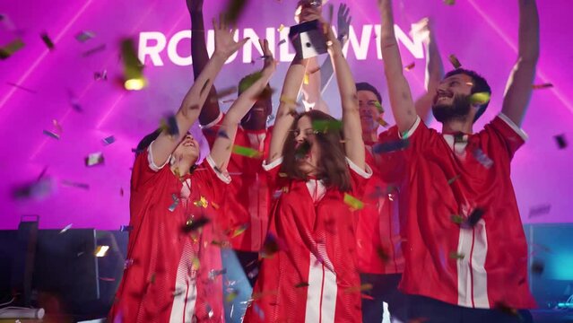 Slow motion, emotions of victory, the red team of esports players rejoices at the victory in the championship, female holds the gold cup in her hands, confetti is flying in the air.