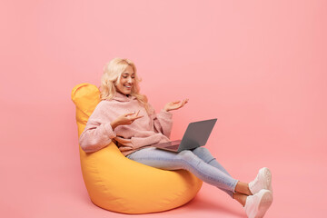 Happy woman making video call on laptop while sitting in beanbag chair, emotionally talking at computer screen - obrazy, fototapety, plakaty