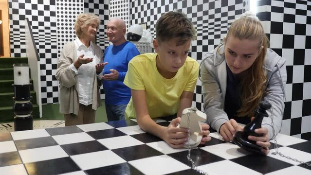 Boy and girl solving chess riddle while standing in escape room. Grandfather and grandmother standing in background and watching them.