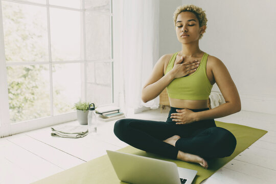 African female yoga teacher having online lesson sitting on green mat in light room showing pranayama techniques, hands on her chest and belly, looking concentrated and focused on body feelings
