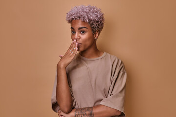 Shocked dazed young African American woman covers mouth with palm hears amazing gossip or said something inappropriate revealing someone else's secret wearing T-shirt stands on brown background