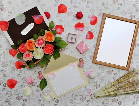 Greeting postcard with free space for writing and blank mock up of photo frame on elegant vintage background with rose petals,box with roses ,gift- box with ring ,2little angels ,fan.Top view