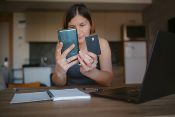 young asian woman at home, remote work at laptop. shopping online at home, lady is holding credit card and using smartphone and computer. Credit card and phone in the focus