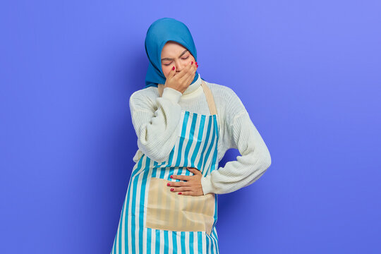 Portrait Of Sick Young Housewife Woman In Hijab And Stiped Apron,  Having Abdominal Pain, Put Your Hand On Your Stomach, Feel Nauseous And Want To Throw Up Isolated On Purple Background