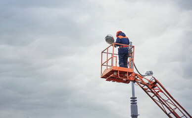 Electrician repairing street light lantern at height. Man in hard hat working in lift bucket, repairing street lamp, led light bulb installation. City street light maintenance, broken bulb replacement