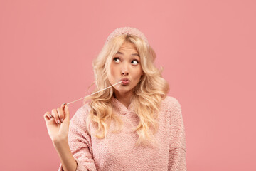 Pretty blonde woman stretching chewing gum from mouth and looking away at free space, pink studio background
