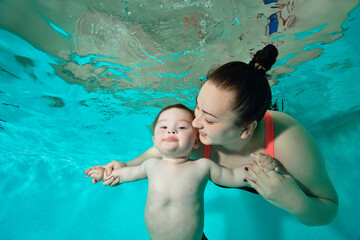 Happy Family: A mother and a child with Down syndrome enjoy swimming underwater in a turquoise water pool. The woman looks at the baby and smiles, holding his hands. Portrait. Horizontal orientation.