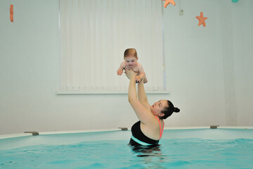 Mom raised and holds a baby with Down syndrome over her head, standing in the water in the pool. An infant swimming coach is engaged with a disabled child in a children's pool. Horizontal orientation.