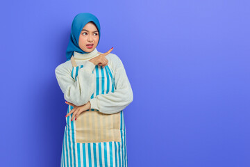 Portrait of annoyed housewife woman in hijab and apron pointing at copy space with finger, looking aside isolated on purple background. People housewife muslim lifestyle concept