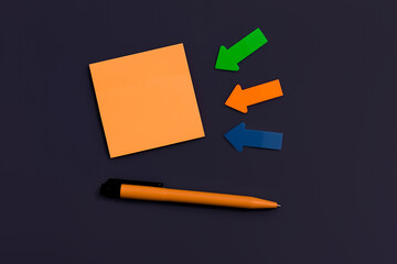 blank orange square sticky note, pen and arrows of paper on dark color background, top view. Blank space for future important events announcements