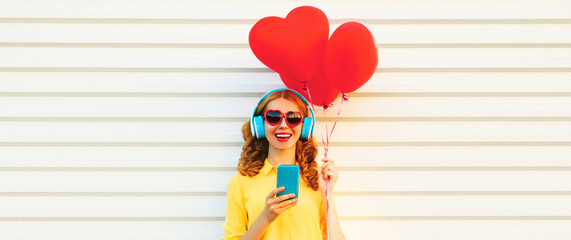 Portrait of happy smiling young woman with smartphone and red heart shaped balloons listening to...