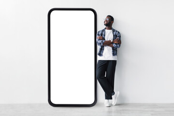 Full body length of black guy standing near huge giant smartphone with empty white screen template,...