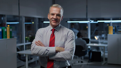 Portrait of smiling mature businessman posing at camera with arms crossed in busy office