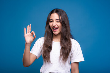 Fototapeta premium Attractive caucasian or arab brunette girl in white t-shirt winking and showing fingers okay gesture isolated on blue studio background.