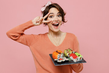 Young cheerful smiling fun woman 20s in casual clothes hold in hand makizushi sushi roll served on black plate traditional japanese food showing victory sign isolated on plain pastel pink background