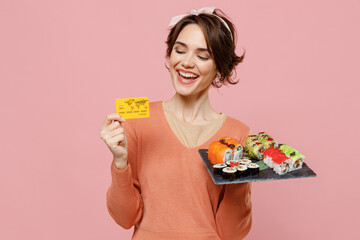 Young satisfied happy woman in casual clothes hold in hand makizushi sushi roll served on black plate traditional japanese food hold in hand credit bank card isolated on plain pastel pink background.