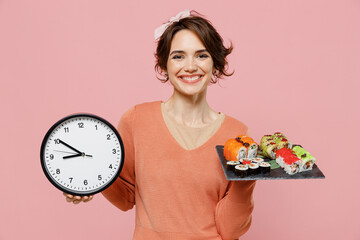 Young smiling happy cheerful caucasian woman 20s wear casual clothes hold in hand makizushi sushi roll served on black plate traditional japanese food clock isolated on plain pastel pink background