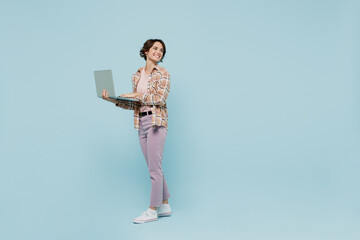 Full body side view young smiling cheerful happy woman 20s wearing brown shirt hold use work on laptop pc computer look aside on area isolated on pastel plain light blue background studio portrait