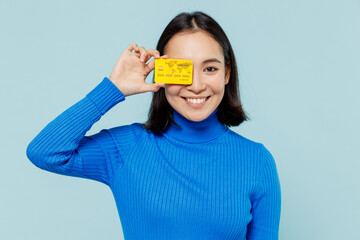 Fascinating fun joyful young woman of Asian ethnicity 20s years old wear blue shirt hold in hand credit bank card cover close hiding eye isolated on plain pastel light blue background studio portrait