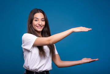 Fototapeta na wymiar Attractive caucasian or arab brunette girl in a white t-shirt showing a big object with her hands on a copy space for advertising isolated on a blue studio background.