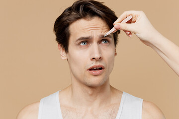 Worried sad young man 20s perfect skin in undershirt hand plucking out hair with tweezers isolated on pastel pastel beige background studio portrait. Skin care healthcare cosmetic procedures concept.