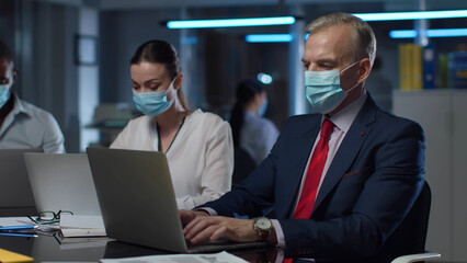 Diverse businesspeople in safety mask work on laptop in modern office at night