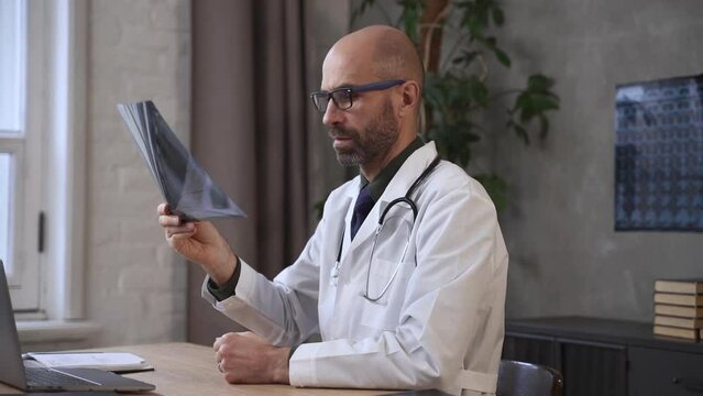Male doctor looking at x-ray and writing while sitting at table with laptop in clinic office spbas. Close view of bearded caucasian man examines shot of lungs and writes in notebook, does diagnostic