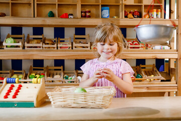 Little preschool girl play with food and grocery wooden toys. Happy active child playing role game as cashier or seller, in wood shop or supermarket. Education, activity for kids.