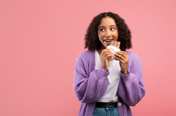 Cheerful millennial African American lady eating chocolate bar over pink studio background, copy...