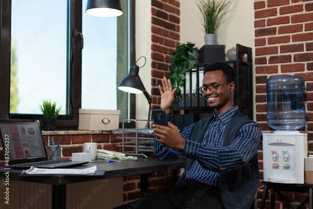 Poster smiling entrepreneur waving hello in video call conference using smartphone and wireless earbuds sit